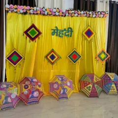 A colorful Rajasthani theme decoration for a Mehndi ceremony. The setup features a bright yellow backdrop, decorative umbrellas, and colorful hanging tassels.