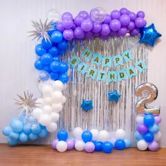 A colorful wall decoration for a second birthday. The setup includes a blue, purple, and white balloon arch, a silver shimmer curtain, a 'Happy Birthday' banner, and a large number '2' foil balloon.