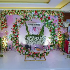 We also create beautiful traditional decorations for events like naming ceremonies. This stage setup features a lush floral arch in pink, white, and red, with a decorated cradle as the centerpiece for the baby.