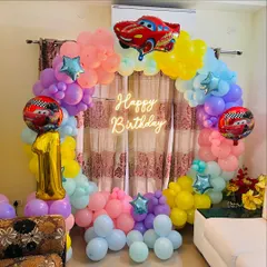 A vibrant "Cars" themed birthday party featuring a full balloon ring in pastel rainbow colors. The decoration is topped with a Lightning McQueen foil balloon and includes a 'Happy Birthday' neon sign.