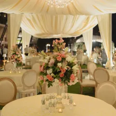 An elegant outdoor dinner setting under a draped canopy with a crystal chandelier. The soft cream and pink floral centerpieces and classic round-back chairs create a sophisticated and intimate atmosphere perfect for a formal corporate dinner.