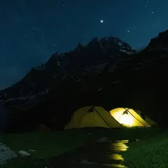 A night camping in the mountains of Kashmir. Tents glow under a sky bright with stars, with the dark, powerful silhouette of the peaks standing guard. The sound of a nearby stream completes the serene experience.