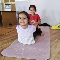 Two young yogis share a smile while helping each other in a partner pose. Our classes for children are designed to be fun, interactive, and build a sense of teamwork.