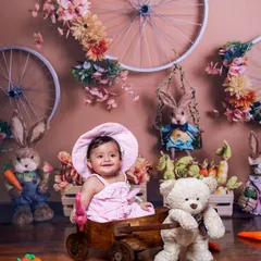 This baby is enjoying a ride in a wooden cart in a rustic, bunny-themed setup. The charming details like the carrots and floral-decorated wheels make this a lovely spring photo.