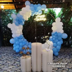 A simple and elegant birthday setup with a circular balloon arch in blue and white. The decor is accented by a neon "Happy Birthday" sign and minimalist white fluted plinths.