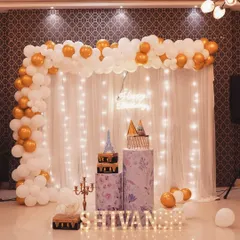 A simple and elegant birthday decoration. This setup features a white and gold balloon arch against a sheer curtain with fairy lights, creating a warm and sophisticated ambiance for an intimate celebration.