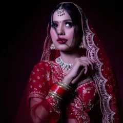 A dramatic, thoughtful pose of the bride looking away from the camera. The lighting sculpts her features and highlights the beautiful embroidery on her red bridal outfit.