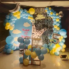 A sky-themed half-birthday decoration for a baby boy. The backdrop features a blue sky with clouds, framed by a yellow and blue balloon arch, a crescent moon foil balloon, and a number '5' foil balloon to mark the 5th-month celebration.