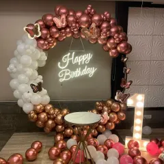 An elegant first birthday setup with a rose gold and white color scheme. The circular balloon arch is accented with butterfly details and surrounds a 'Happy Birthday' neon sign, with a marquee number '1' on the side.