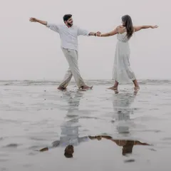 A playful, dancing moment on a wet beach during a monsoon pre-wedding shoot. The reflection in the water and their joyful movement create a lighthearted and romantic image.
