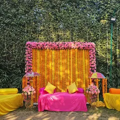 This vibrant mehendi backdrop combines traditional marigold strings with a border of bright pink flowers. The comfortable pink seating provides a perfect spot for the bride during her mehendi application.