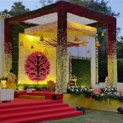 A vibrant and traditional floral mandap. The structure is covered in a gradient of red, yellow, and white flowers, with a Ganesha motif made of pink carnations as the centerpiece, creating a colorful and auspicious setting.