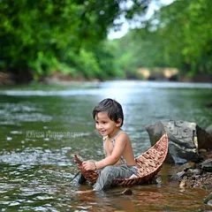 A wider shot of the boy in the boat, capturing the beautiful, lush green backdrop of the river and trees.