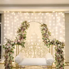 This is a classic and elegant seating arrangement for an engagement or roka. The white wrought-iron bench is framed by beautiful pink and white floral arrangements and a backdrop of warm fairy lights.