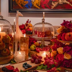 The tablescape is a study in texture and color, with the rich reds of the flowers and pomegranates contrasting with the natural wood and patterned tablecloth. The artwork in the background adds another layer of sophistication.