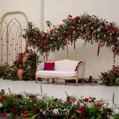 A regal reception stage designed with a stunning arch of deep red florals. The classic white sofa and terracotta pots add a touch of vintage elegance to the grand setting.