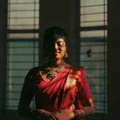 An artistic and moody portrait of a bride in a red saree. The play of shadow and light from the window creates a dramatic, vintage feel, highlighting her silhouette and the richness of her attire.