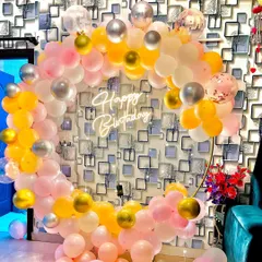 A cheerful and bright balloon ring against a patterned wallpaper. The mix of pink, yellow, white, and silver balloons creates a fun and festive photo corner.