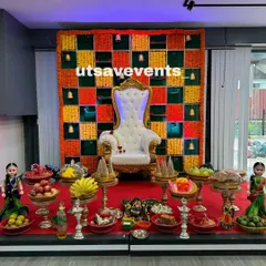 A complete baby shower setup with a marigold checkerboard backdrop and a stage filled with traditional offerings and dolls. The high-back throne chair adds a regal touch.