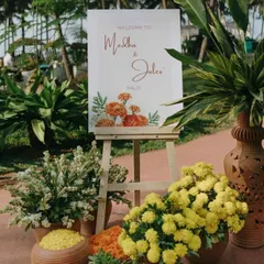 A simple and elegant welcome sign for a Haldi ceremony, surrounded by pots of marigolds and chrysanthemums.