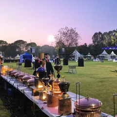 An expansive outdoor catering setup at dusk. The beautifully lit buffet line with ornate copper chafing dishes and decorative lamps creates a magical atmosphere as day turns to night.