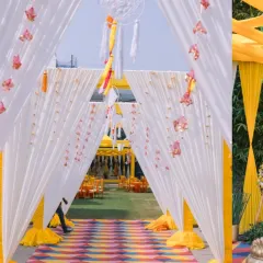 A cheerful and bright entrance for a Haldi ceremony, with white and yellow drapes, dreamcatchers, and colorful floor mats to welcome guests.
