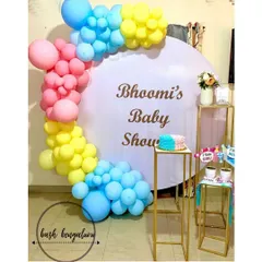 A bright and cheerful baby shower for Bhoomi, using a vibrant mix of pink, yellow, and blue balloons on a clean white circular backdrop. The setup includes gold pedestals for the cake and props.