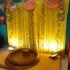 A simple and traditional Haldi backdrop for a home function. The setup includes a bright yellow curtain, decorative umbrellas, and a low wooden stool (chowki) for the ceremony.
