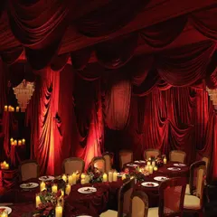 An immersive dining experience designed for an intimate engagement. The entire room is draped in red velvet, with tables set for a royal feast, illuminated by candlelight and grand chandeliers.