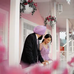 A beautifully soft and romantic shot of Manpreet and Simranpreet on the porch of a pastel pink house set. The foreground flowers add depth and a dreamy quality to the photo.