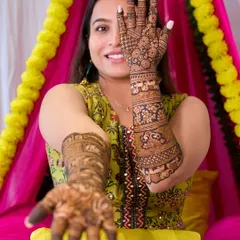 A classic bridal portrait for a Mehndi ceremony, where the bride playfully shows off her intricate henna designs. The shot is framed to highlight both her joyful expression and the detailed artistry on her hands and arms.