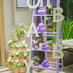 This 'Oh Baby' ladder shelf is a charming and versatile decor piece. Dressed in a soft lavender hue and adorned with small floral pots and balloon accents, it serves as a perfect welcome element or a creative photo prop for your baby shower.