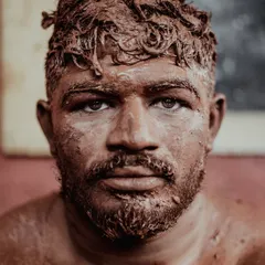An intense close-up portrait of a wrestler, his face and hair caked with the reddish-brown soil of the taleem. His direct gaze conveys the focus and determination required for the sport.