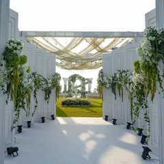 A sun-drenched view through the grand white gates of a Goa wedding. The path opens up to a lush green lawn, with a floral arch and fountain in the distance, setting the scene for a romantic outdoor ceremony.