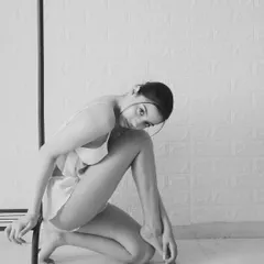 A simple, minimalist black and white shot. The model's pose is fluid and graceful, using a clothing rack as a prop to create interesting lines and shapes.