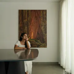 This shot captures the serene ambiance of the dining space. The custom-designed table with its unique leg structure is a statement piece, complemented by the soft light from the sheer curtains and the organic feel of the artwork.