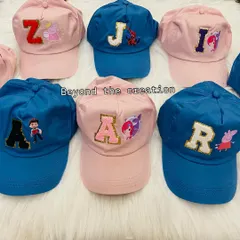An overhead view of several personalized caps ready for a party. Each cap, in either blue or pink, features a unique combination of an initial and a character patch, from Peppa Pig to superheroes.