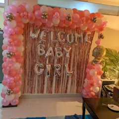 A beautiful 'Welcome Baby Girl' decoration for a home celebration, featuring a full balloon arch in shades of pink and white against a rose gold fringe curtain.