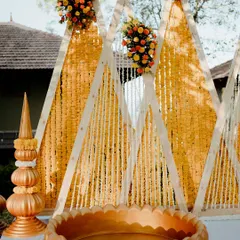 A modern take on Haldi decor, this setup features geometric triangle backdrops draped with marigold and tuberose garlands. The large golden urli in the foreground is ready for the ceremony.