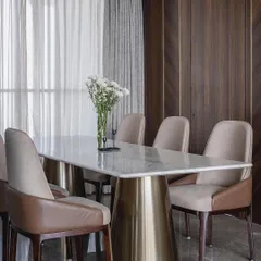 The dining area in the 'Aria Residence' exudes contemporary elegance. A white marble tabletop rests on a sculptural brass base, surrounded by chairs upholstered in leather and fabric. The dark veneer wall adds a rich, warm backdrop.