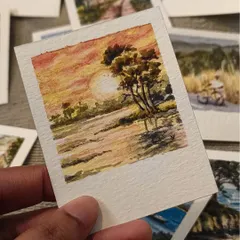 A warm, golden sunset over the water, painted in watercolor on a miniature Polaroid. The reflection of the sun and the silhouette of the tree create a peaceful, dreamy atmosphere.