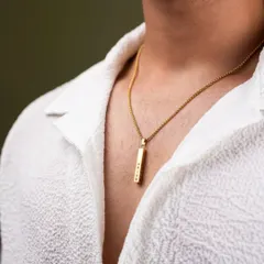 This lifestyle shot shows how a piece of jewelry complements personal style. The focus is on the gold pendant against a simple white shirt, demonstrating the product in a natural, wearable context for a men's accessory brand.