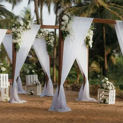 An elegant and minimalist beach wedding mandap. The simple wooden structure is draped with white fabric and adorned with classic white floral arrangements, letting the beauty of the ocean view shine.