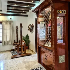 This image showcases a beautifully crafted pooja cabinet made from polished dark wood with detailed carvings and latticework on the glass doors. Positioned in the living area, it serves as both a spiritual corner and a piece of fine furniture.