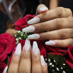 A close-up of our intricate bridal nail extensions, featuring white sculpted patterns over a pearlescent base, accented with subtle rhinestones. This design is set against a bouquet of red roses, highlighting its romantic and celebratory feel.