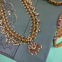 A traditional pearl necklace set, also known as a moti haar. This design features delicate pearls clustered with ruby and emerald colored stones, offering a timeless and elegant look for any occasion.
