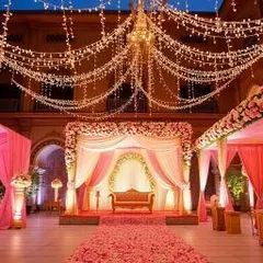 A classic palace courtyard wedding setup. The decor features a pink and white theme with a floral stage, draped canopies, and a ceiling of fairy lights, creating a royal and romantic atmosphere.