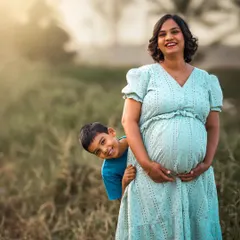 A joyful outdoor family portrait where a young boy playfully peeks out from behind his pregnant mother. The golden hour sunlight adds a warm, happy glow to this candid moment.