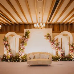 A clean and elegant stage design for an engagement ceremony. The backdrop features modern white arches adorned with delicate pink and white floral arrangements, creating a soft and romantic focal point.