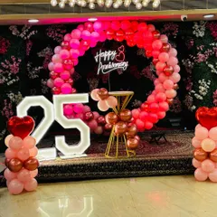 A beautiful 25th anniversary stage setup. It features a large balloon ring in red and pink, light-up marquee numbers, and a 'Happy Anniversary' neon sign against a floral backdrop.